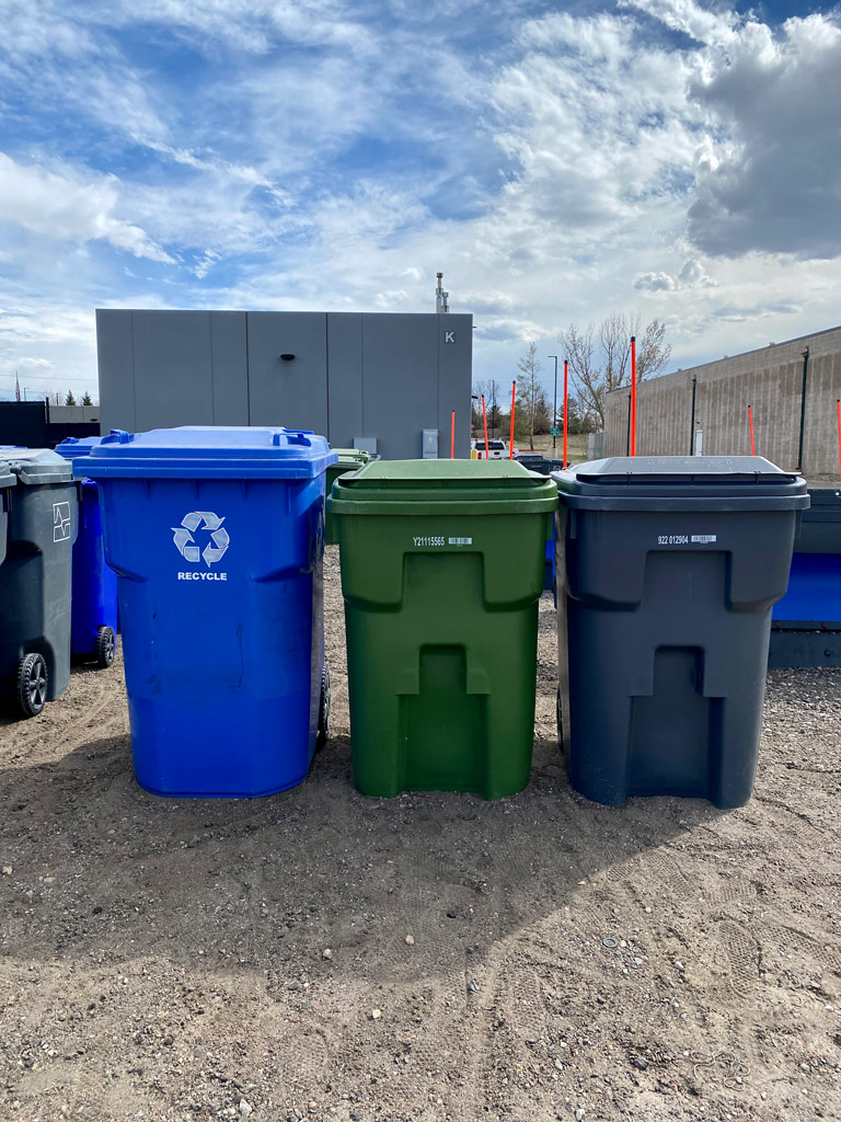 Three different types of solid waste bins are lined in a row, each a different color. One for recycling, yard waste and trash.