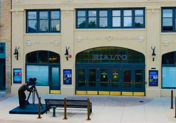 An aerial photograph of the Rialto Theater Center entrance. There is a sculpture of a person looking through a film camera and a bench outside the entrance.